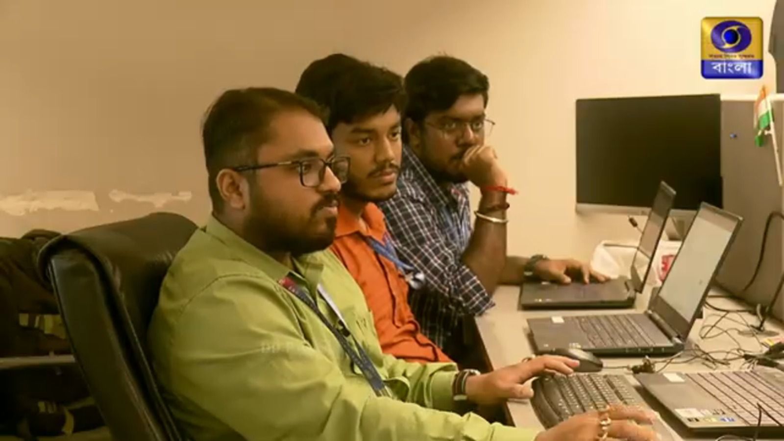 Still from the broadcast: three colleagues at a shared office desk working on laptops, a small Indian flag standing on the desk behind them, with the DD Bangla channel logo in the upper right of frame.